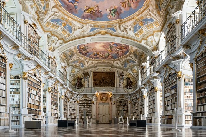 Interior grandioso de biblioteca com tetos ornamentados e estantes altas repletas de livros em uma atmosfera mágica.