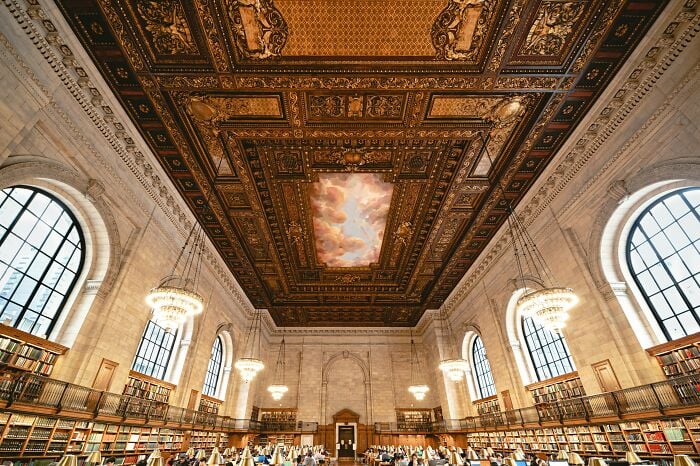 Interior grandioso de biblioteca com teto ornamentado, grandes janelas em arco, lustres e fileiras de estantes cheias de livros. Interior grandioso de biblioteca com teto ornamentado, grandes janelas em arco, lustres e fileiras de estantes cheias de livros.