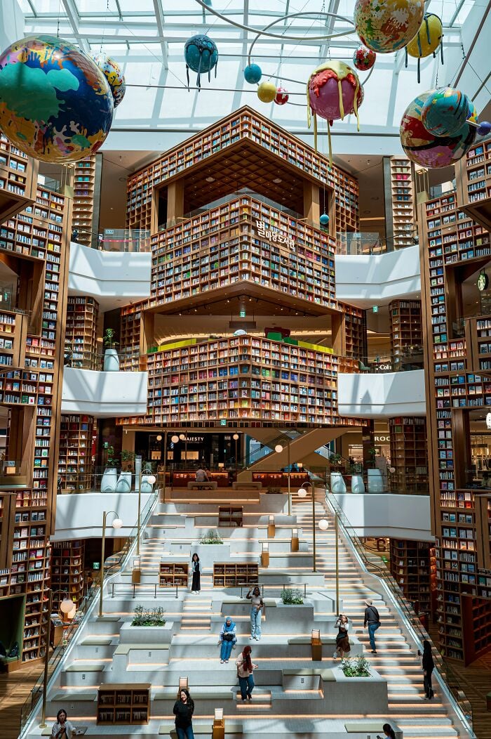 Interior grandioso de biblioteca com estantes imponentes, grande claraboia e esculturas coloridas de planetas penduradas no teto. Interior grandioso de biblioteca com estantes imponentes, grande claraboia e esculturas coloridas de planetas penduradas no teto.
