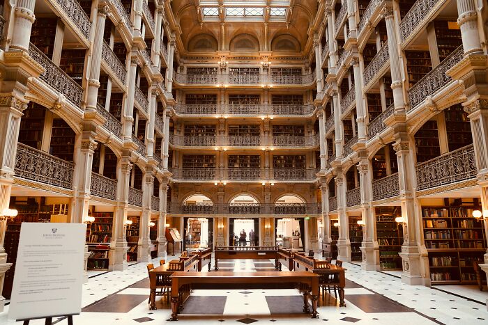Grandiose library interior with multiple levels of bookshelves, ornate columns, and a spacious reading area with wooden tables and chairs. Grandiose library interior with multiple levels of bookshelves, ornate columns, and a spacious reading area with wooden tables and chairs.